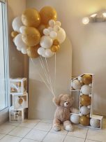 Un ours en peluche tient des ballons dorés et blancs près du chiffre 1.