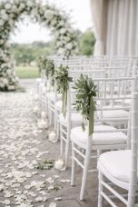 Chaises blanches décorées de verdure, allée de pétales et arceau floral en arrière-plan.